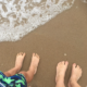 two pairs of bare feet standing on sand and ocean water